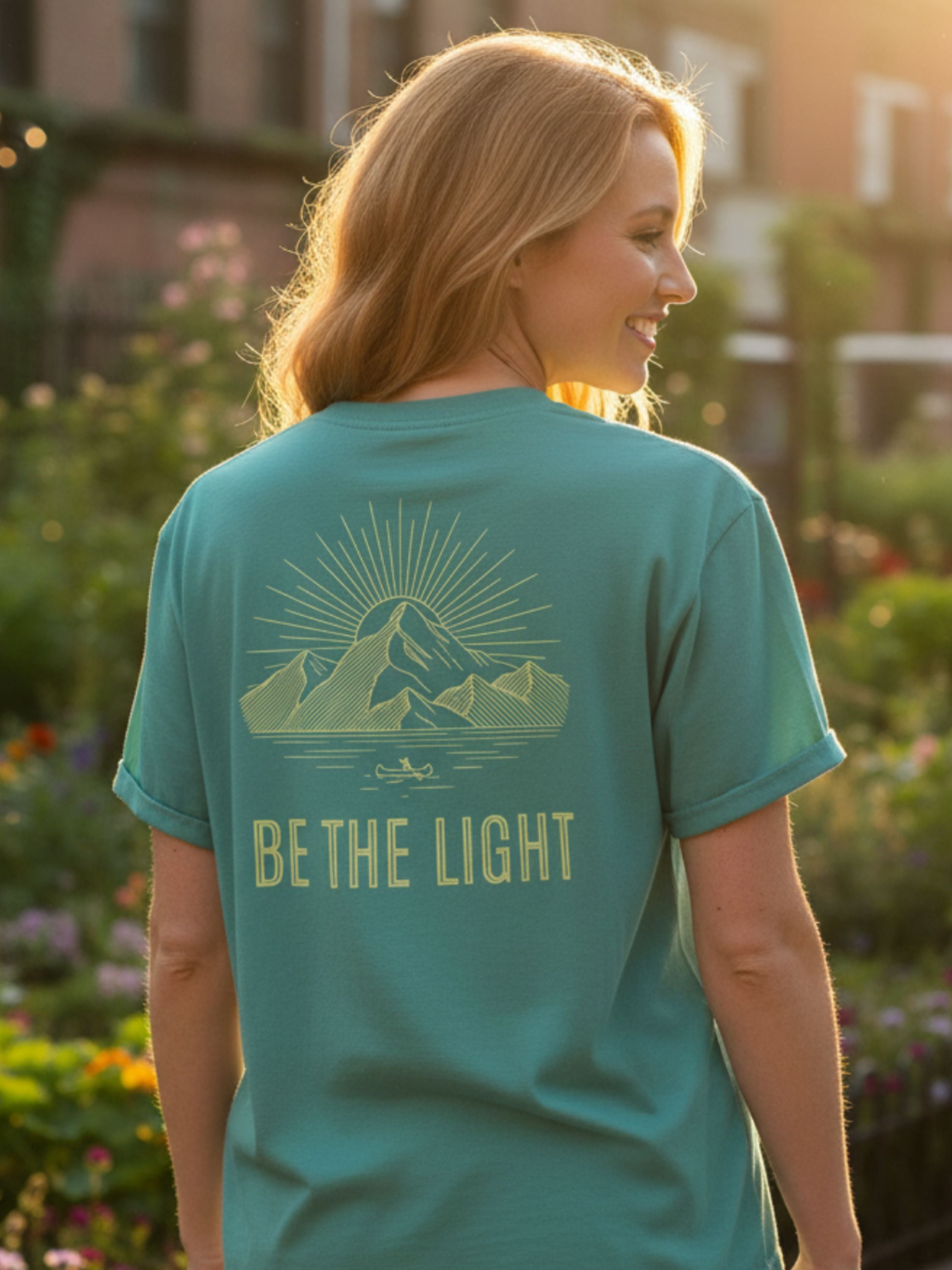 Woman wearing a teal t-shirt with 'Be the Light' message outdoors.