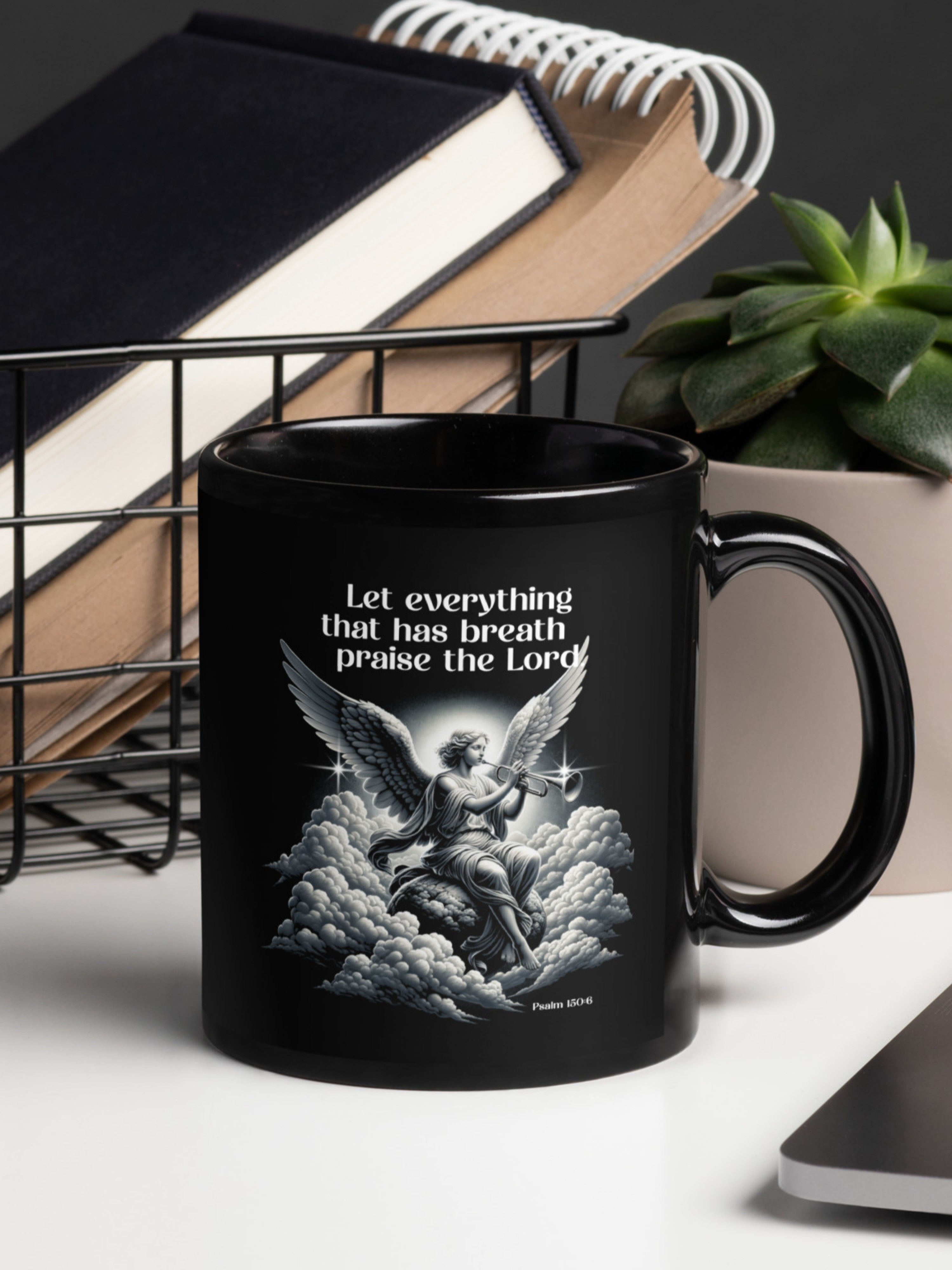 Black mug with angel design and praise text on a desk with books and a plant.