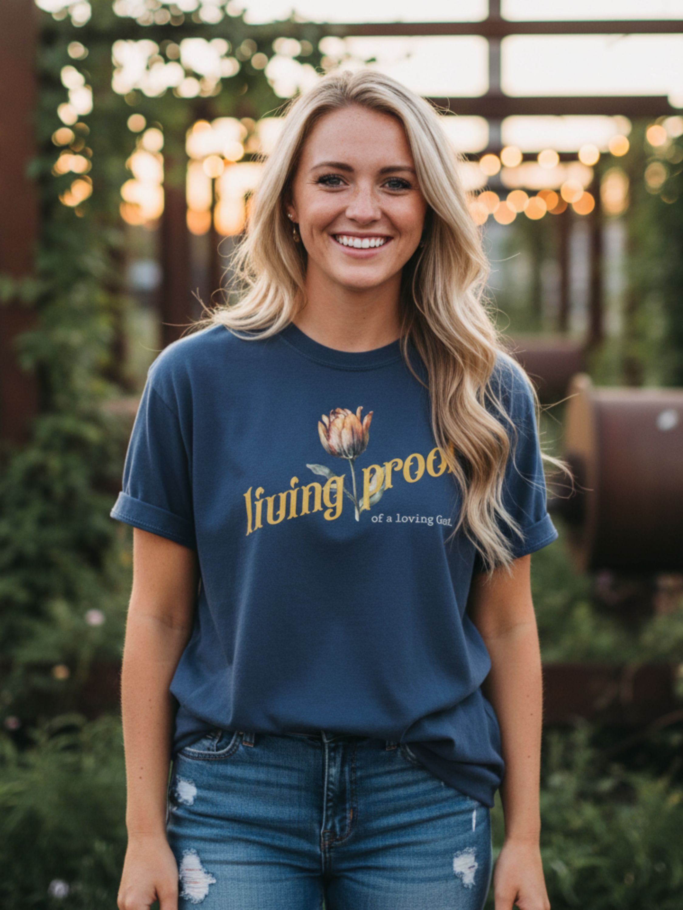 Woman wearing a blue t-shirt with a logo and text, standing outdoors with blurred lights in the background.